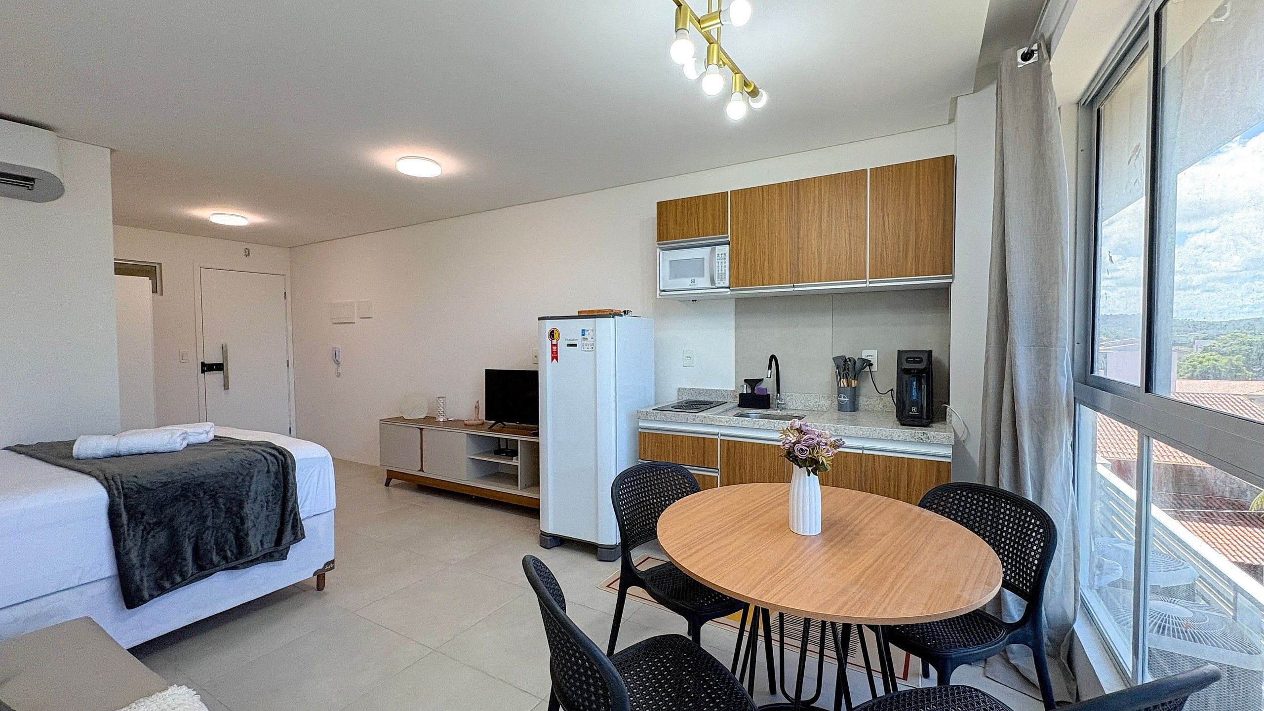 A compact hotel room with a bed, a small kitchenette, a dining area with a table and chairs, and a window providing a view.