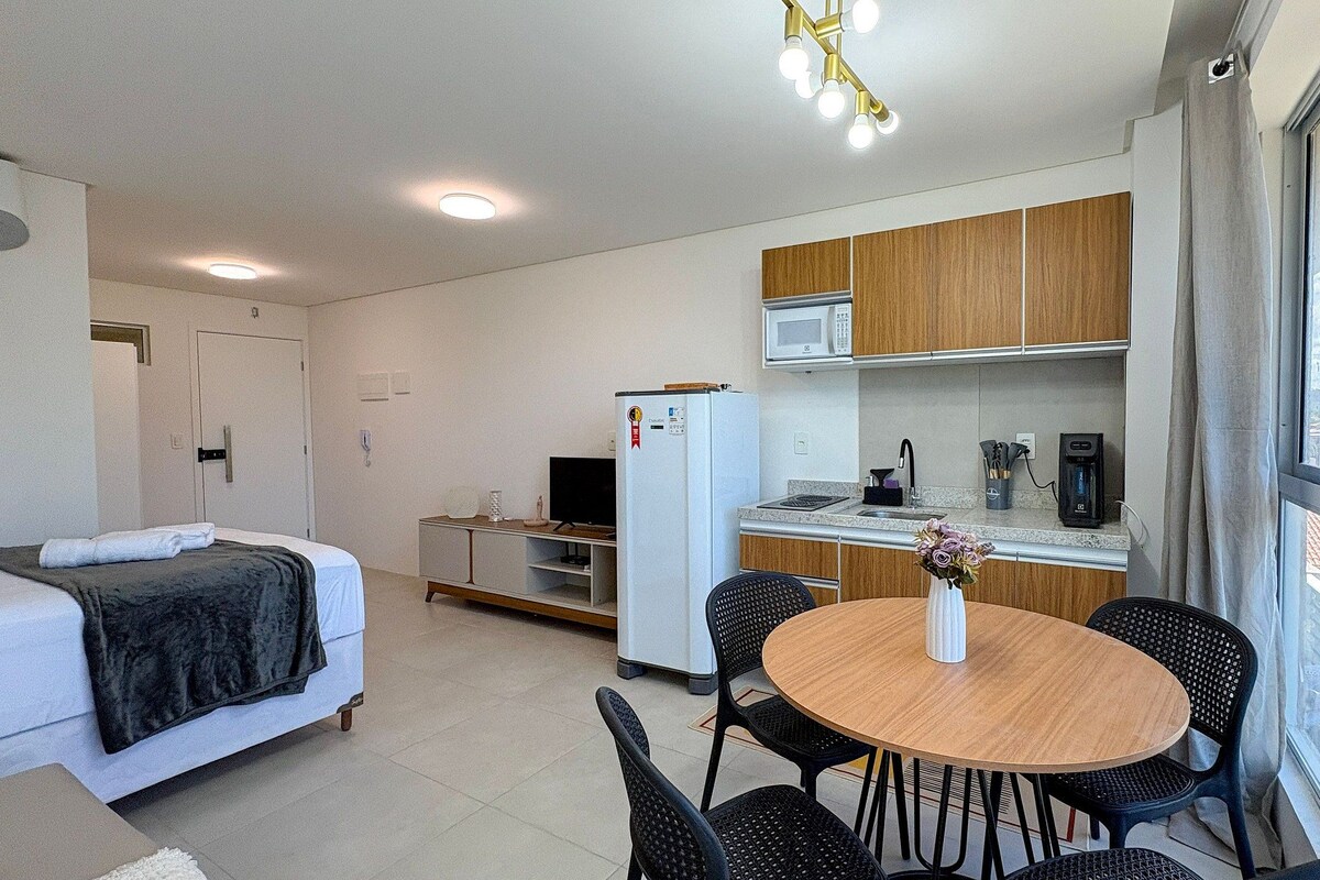 A compact hotel room with a bed, a small kitchenette, a dining area with a table and chairs, and a window providing a view.