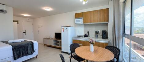 A compact hotel room with a bed, a small kitchenette, a dining area with a table and chairs, and a window providing a view.