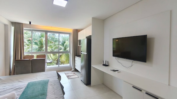 A modern hotel room with a bed, a TV, a refrigerator, and a window with a view of palm trees. - Mila Rooftop - Praia de Tamandaré (Tamandaré)
