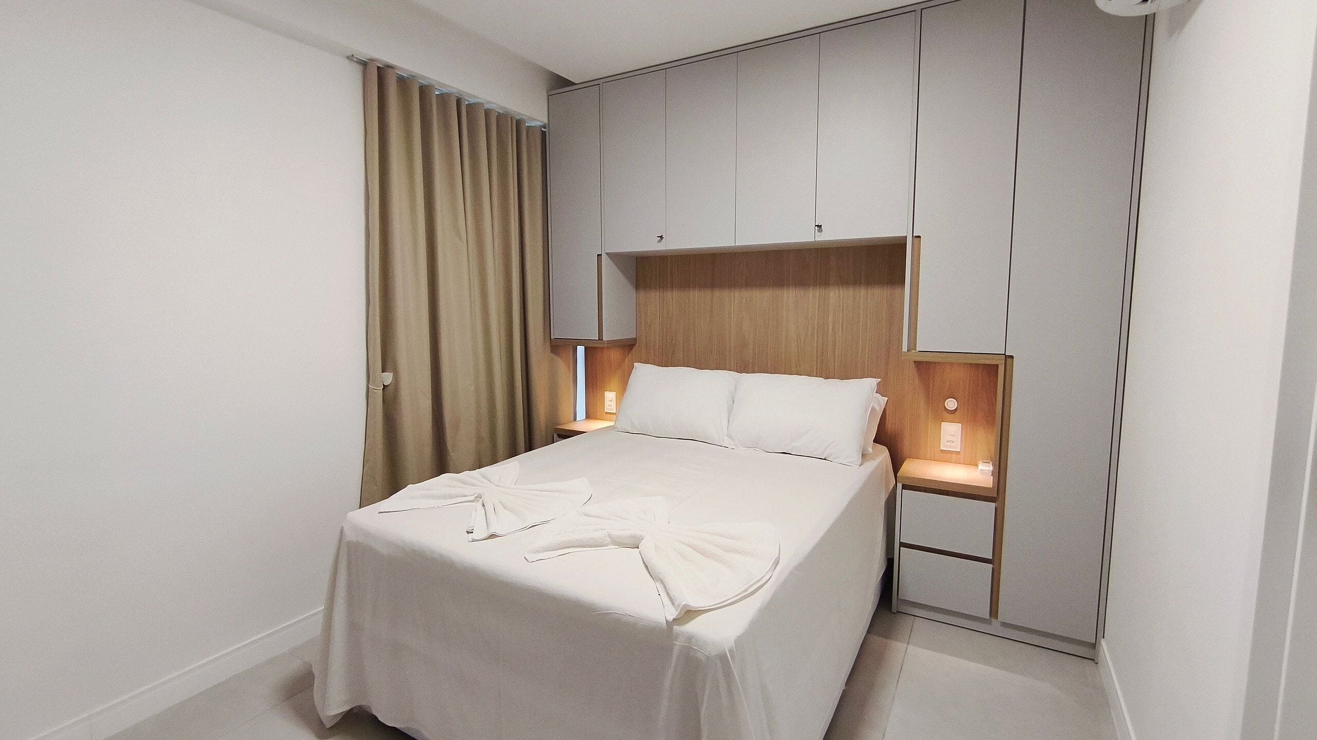 A hotel room with a bed, curtains, a nightstand, and a wall unit with storage.