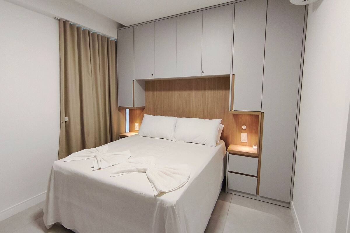 A hotel room with a bed, curtains, a nightstand, and a wall unit with storage.
