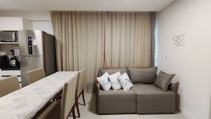 Un espacio habitable compacto con kitchenette, un sofá con cojines, una mesa de comedor y una cortina.