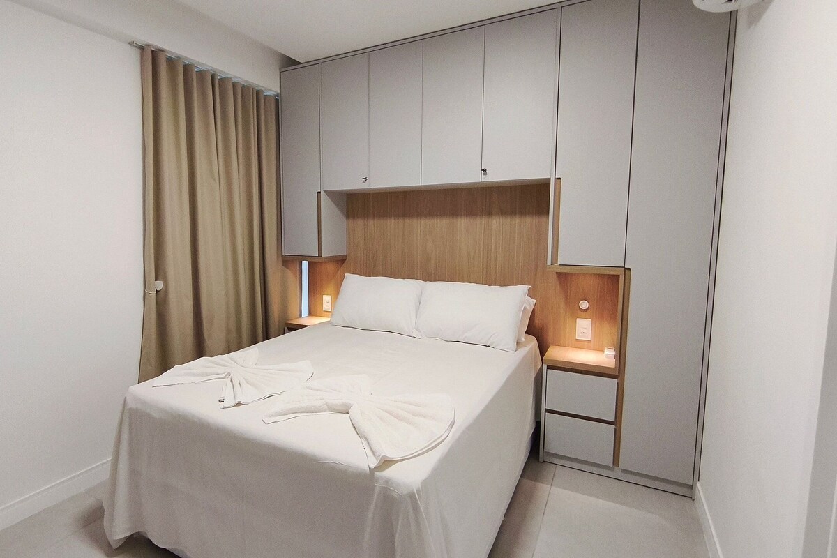 A hotel room with a bed, curtains, a nightstand, and a wall unit with storage.