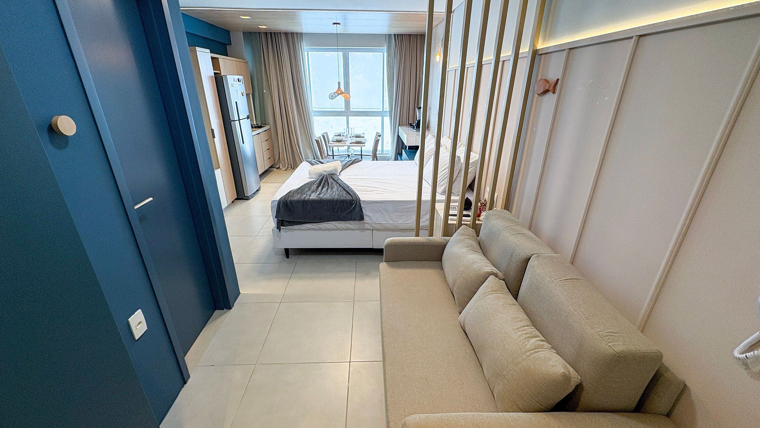 A modern hotel room with a bed, a sofa, a dining area, and a view of the outdoors.