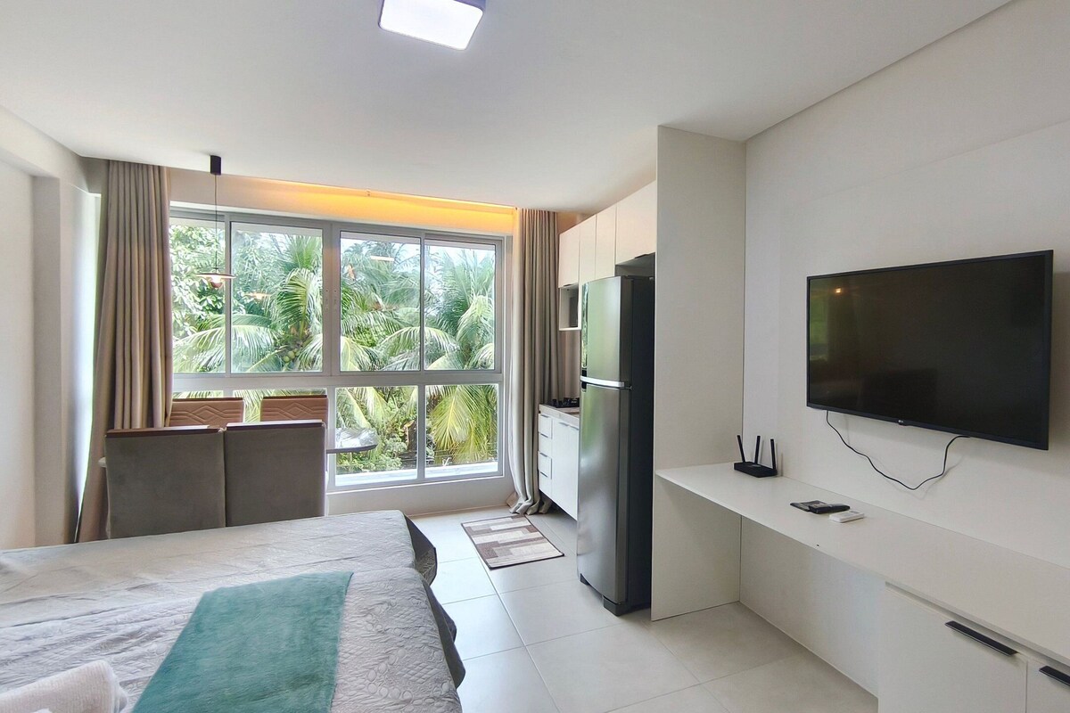 A modern hotel room with a bed, a TV, a refrigerator, and a window with a view of palm trees.