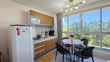 A compact kitchen with a refrigerator, microwave, and sink. There's a dining area with a table and chairs, and a large window offering a view of palm trees.