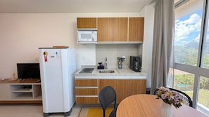 A compact kitchen with wooden cabinets, a microwave, a refrigerator, a coffee maker, and a dining area with a table and chairs. - Mila Rooftop - Praia de Tamandaré (Tamandaré)