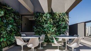A rooftop terrace with a round table and white chairs, surrounded by lush green plants.