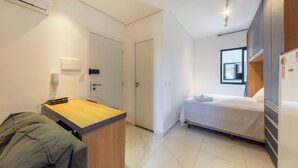 A modern hotel room with a bed, a desk, and a chair. There is a window providing natural light and a door leading to another room.