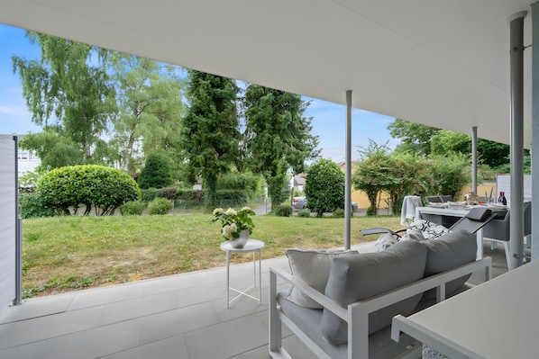 A covered outdoor seating area with a white table, a sofa with cushions, and a small round table with a plant.