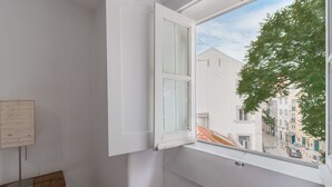 A room with a white wall, a wooden bench, a white door, and a window with a view of buildings and a tree.
