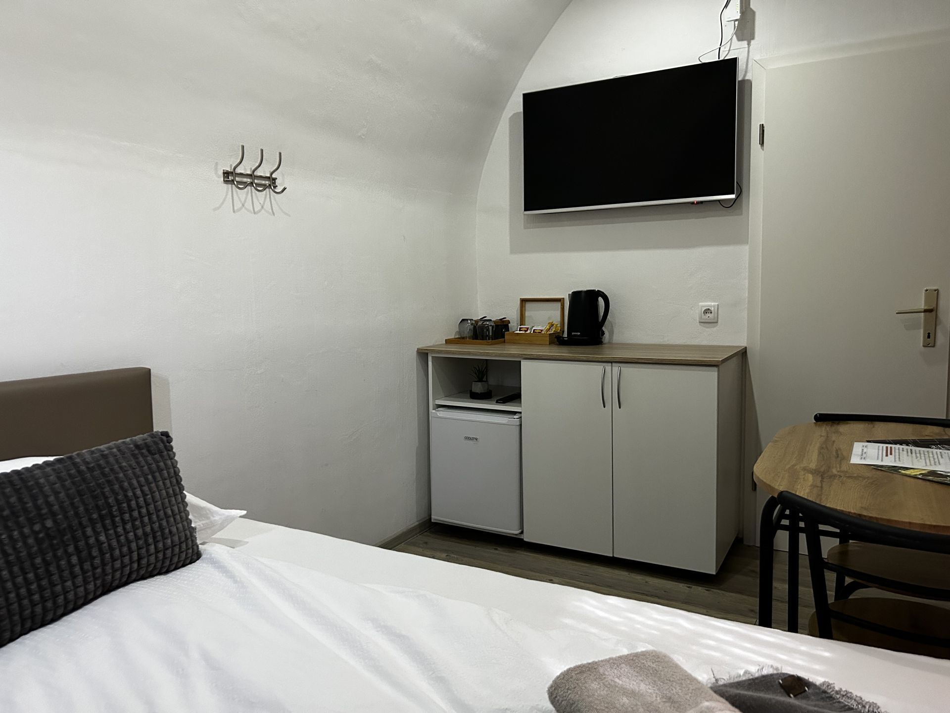Habitación de hotel pequeña con cama, kitchenette, televisión de pantalla plana en la pared y mesa de comedor.