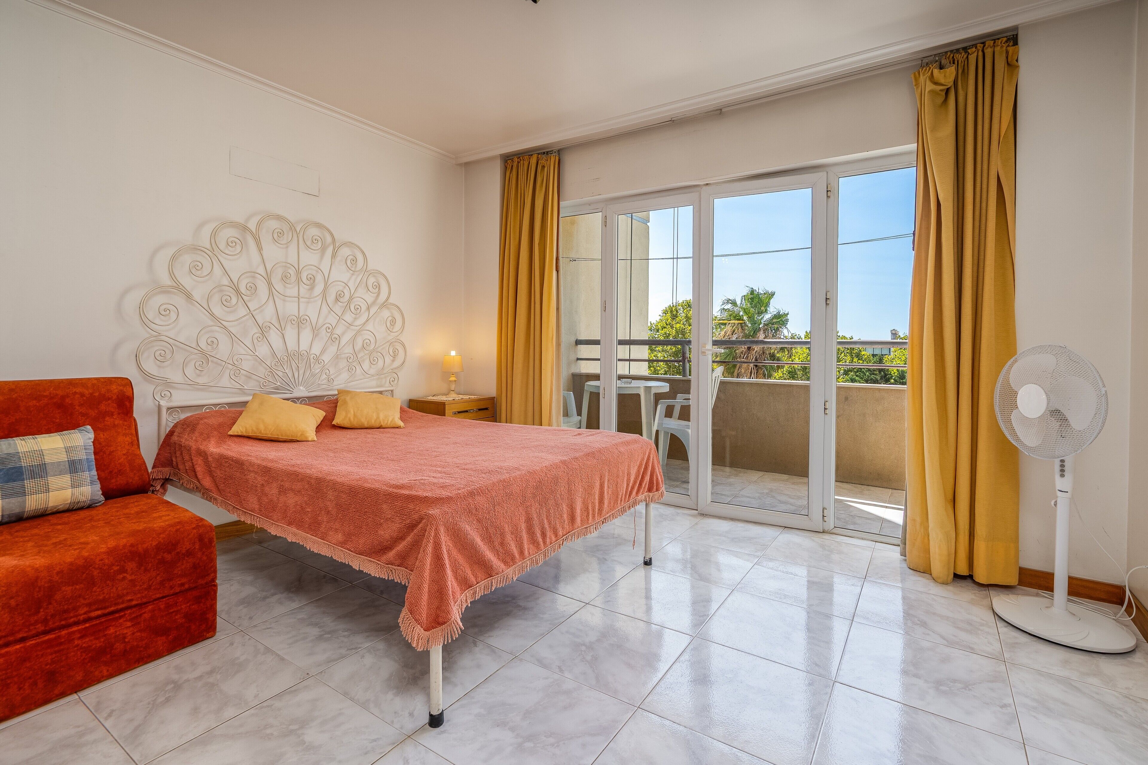 A bedroom with a bed, a red sofa, a standing fan, and a balcony with yellow curtains.