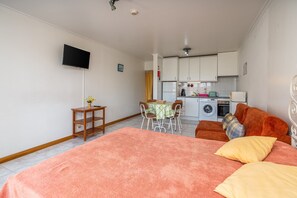 Ein Schlafzimmer mit einem Bett, einem Fernseher, einem kleinen Tisch mit einer Vase und einer Küchenzeile im Hintergrund.