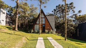 A triangular wooden cabin with a visible door and windows, surrounded by trees and a grassy area with a concrete path leading to it. - RJF - Comfortable Chalets in Urubici (Urubici)