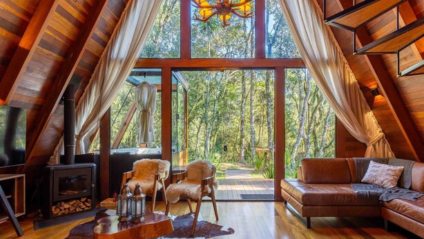 A cozy, wooden interior with a large glass door leading to a forest view, a fireplace, and a comfortable seating area. - RJF - Comfortable Chalets in Urubici (Urubici)