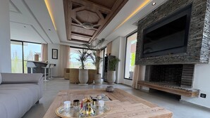 A modern living room with a large wooden coffee table, a stone fireplace, and a flat-screen TV mounted on the wall. - New 480 m² villa with service 10 mn from center (Marrakech)