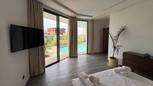 A hotel room with a large bed, a flat-screen TV mounted on the wall, a wooden dresser, a potted plant, and a sliding glass door leading to a pool area. - New 480 m² villa with service 10 mn from center (Marrakech)
