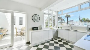 Una cocina moderna con armarios blancos, piso a cuadros blanco y negro, un ventanal con vista a palmeras y un reloj de pared.