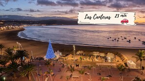 Vista de la playa con un gran árbol de Navidad azul, una carpa blanca y una zona rocosa con gente.