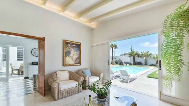Amplia sala de estar con una gran mesa de vidrio, muebles de mimbre, un cuadro y vistas a una piscina y jardín.