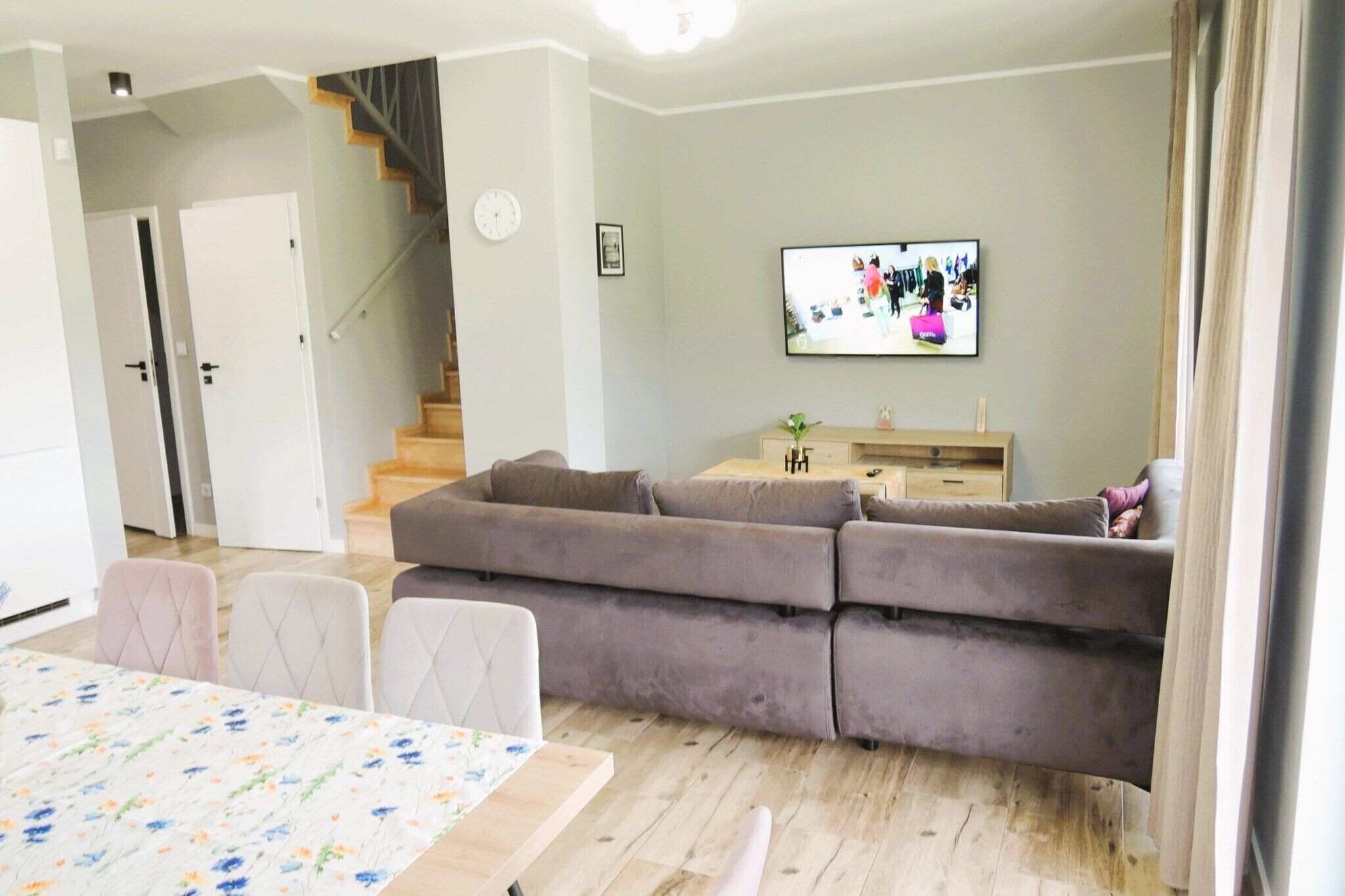 A modern living room with a grey sofa, a dining table with chairs, a staircase, and a flat-screen TV on the wall.