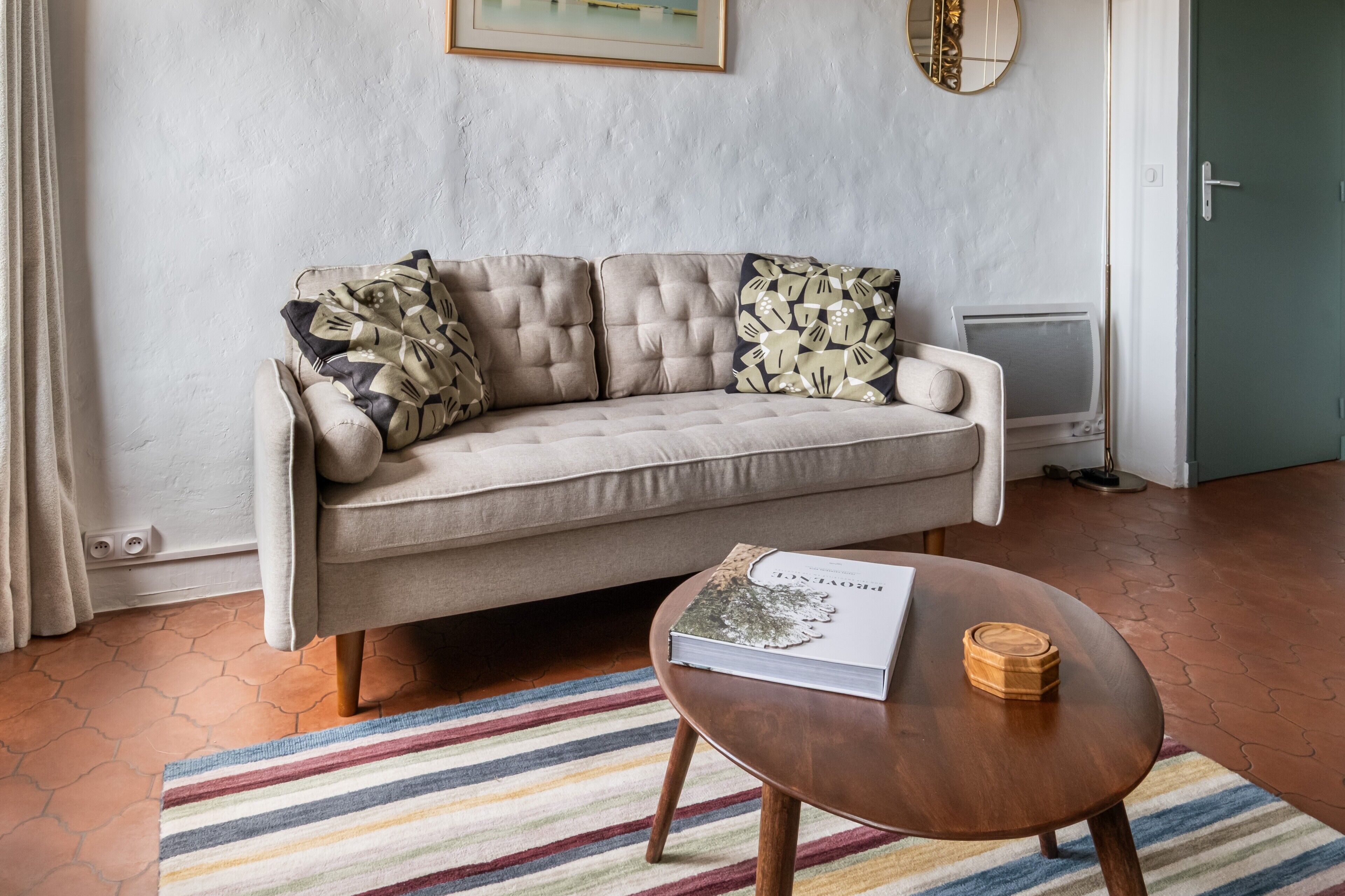 A beige sofa with patterned cushions, a round wooden coffee table with a book and a coaster, a striped rug, and a framed picture on the wall.