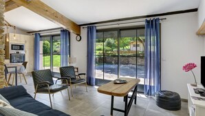 Sala de estar con sofá, sillas, una mesa de madera, una zona de cocina y ventanas amplias con cortinas.