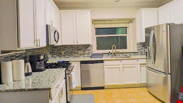 Una cocina con armarios blancos, un refrigerador de acero inoxidable, un lavavajillas, una estufa y una ventana con persianas.
