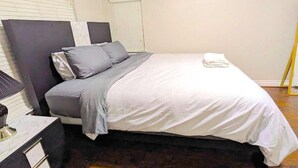 Una cama bien hecha con ropa de cama blanca y gris, una mesita de noche con una lámpara y una ventana con persianas.