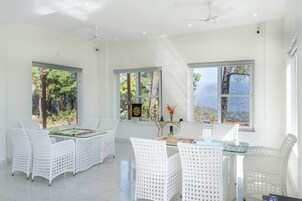 Una habitación luminosa y amplia con muebles de mimbre blanco, ventanas grandes con vistas a árboles y montañas, y un ventilador de techo.