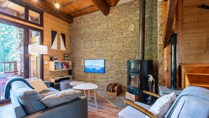 A cozy cabin interior with a stone wall, wooden beams, a fireplace, a sofa, a table, and a bookshelf.