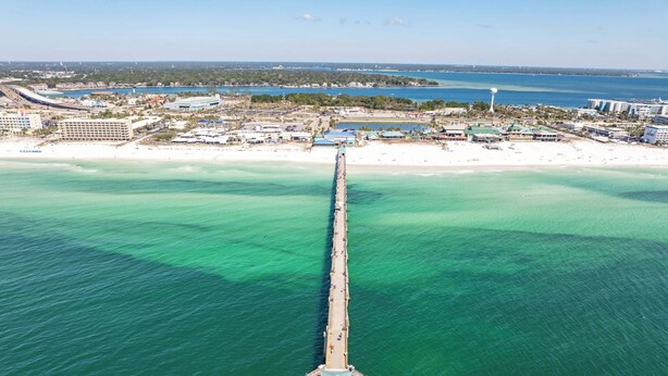 Vista aérea de un muelle que se extiende hacia aguas turquesas, rodeado por una ciudad costera con edificios y una playa.