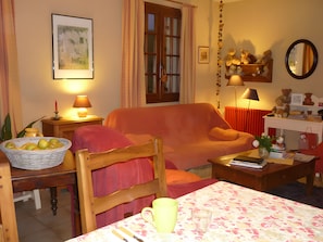 A cozy living room with a wooden sofa, a dining table with a basket of fruit, a lamp, and a mirror on the wall.