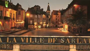 En historisk plads med traditionel europæisk arkitektur, et markant skilt med teksten 'A LA VILLE DF SARLAT' og en belagt grund.