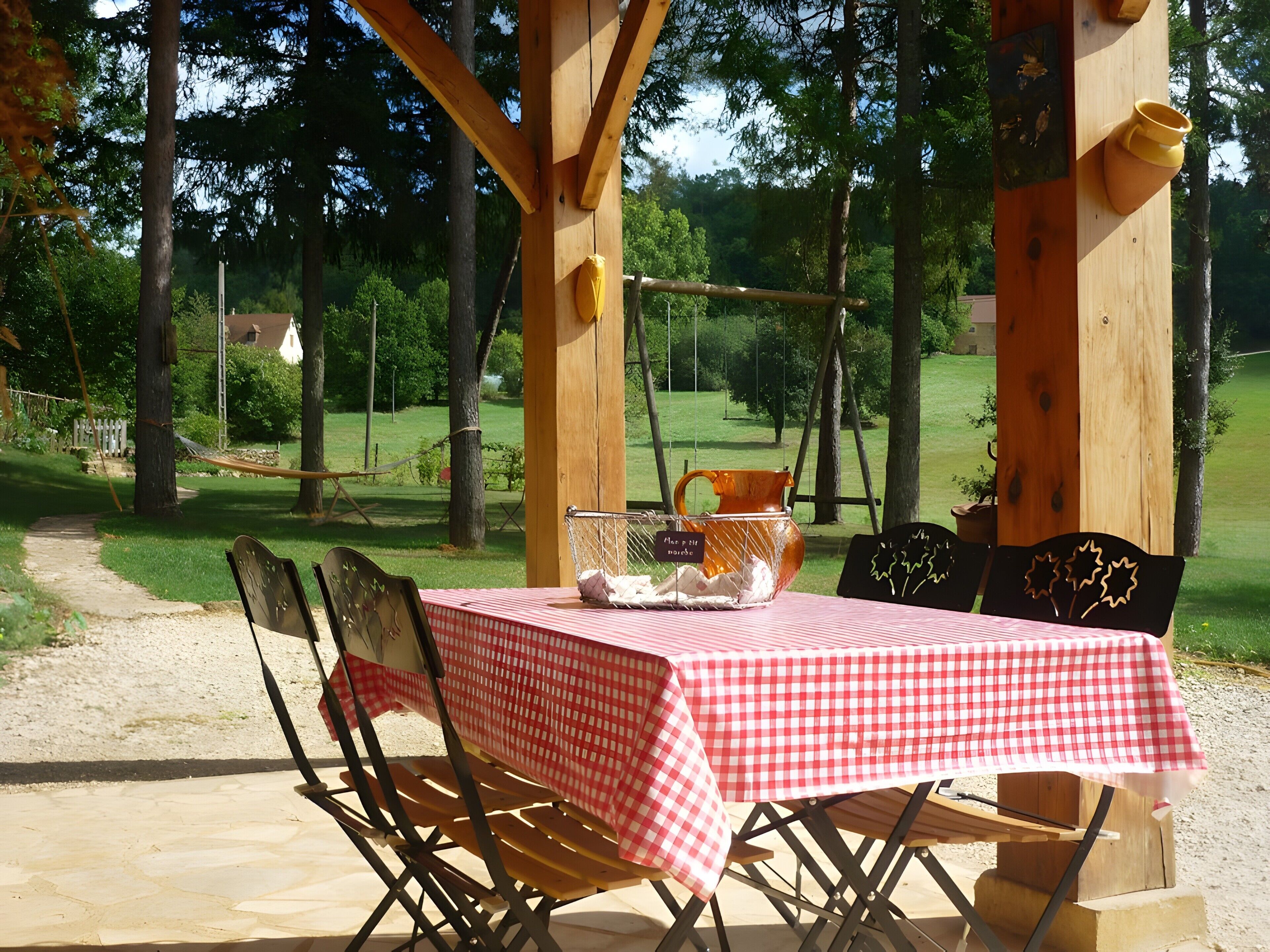 An outdoor wooden pergola with a checkered tablecloth-covered table and chairs, surrounded by trees and a grassy area.