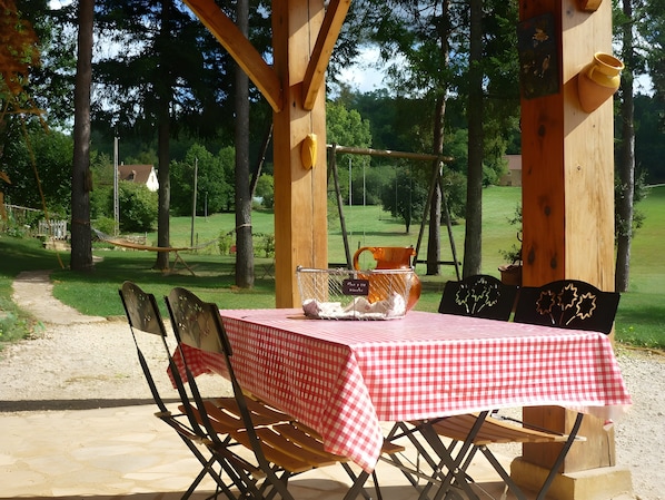 An outdoor wooden pergola with a checkered tablecloth-covered table and chairs, surrounded by trees and a grassy area.