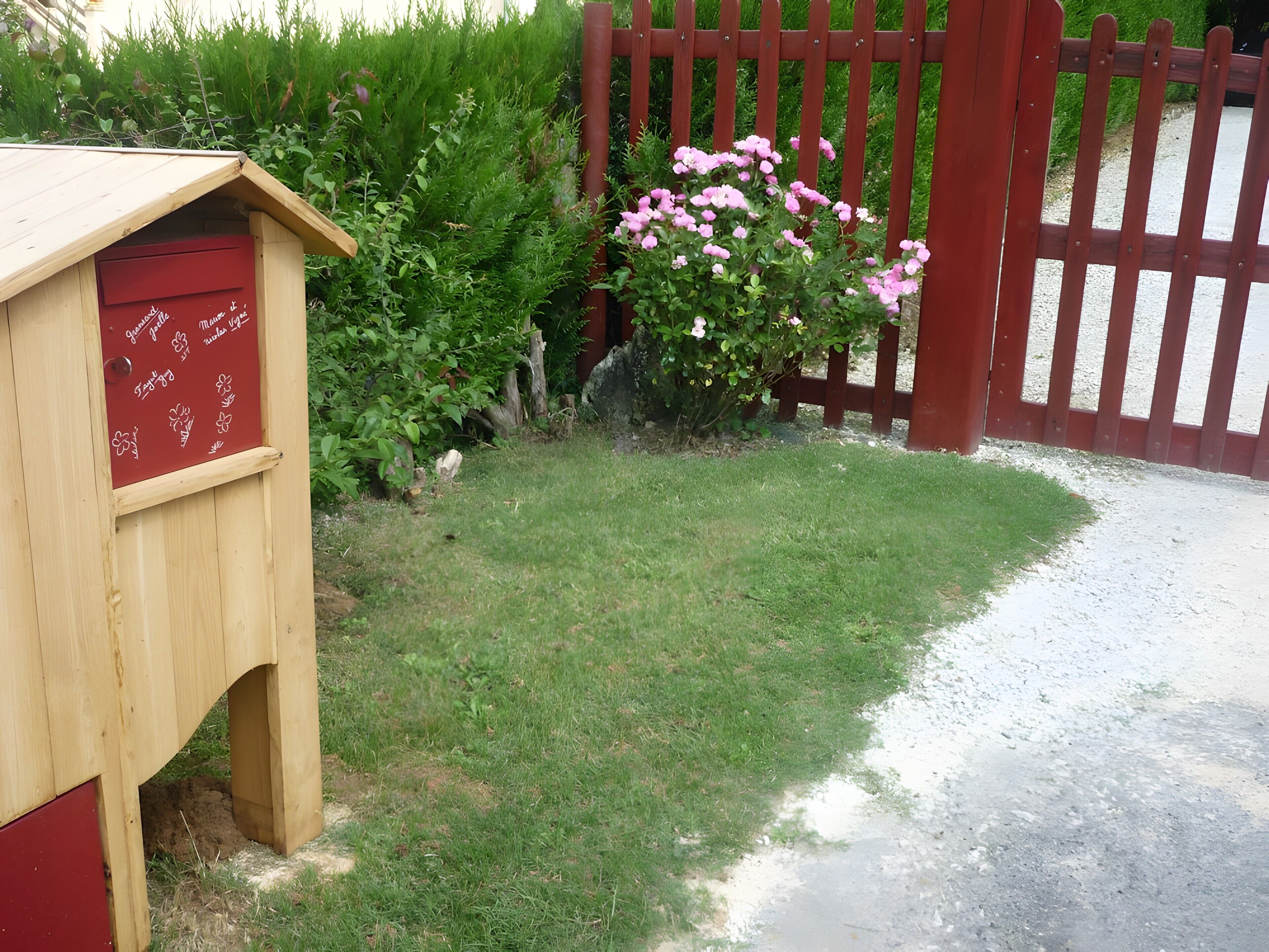 Une structure en bois avec une porte rouge, entourée d’herbe verte et d’une clôture rouge.