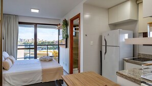 Een moderne hotelkamer met een bed, een houten tafel, een kitchenette met een koelkast, en uitzicht op de stad door grote ramen.