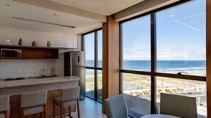 A modern kitchen with a large window overlooking the ocean, a dining area with chairs, and a refrigerator. - Sea View 2Bedroom Apartment on Salvador’s Shore (Salvador)