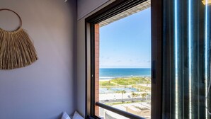 A room with a large window offering a view of the ocean and a coastal road. - Sea View 2Bedroom Apartment on Salvador’s Shore (Salvador)
