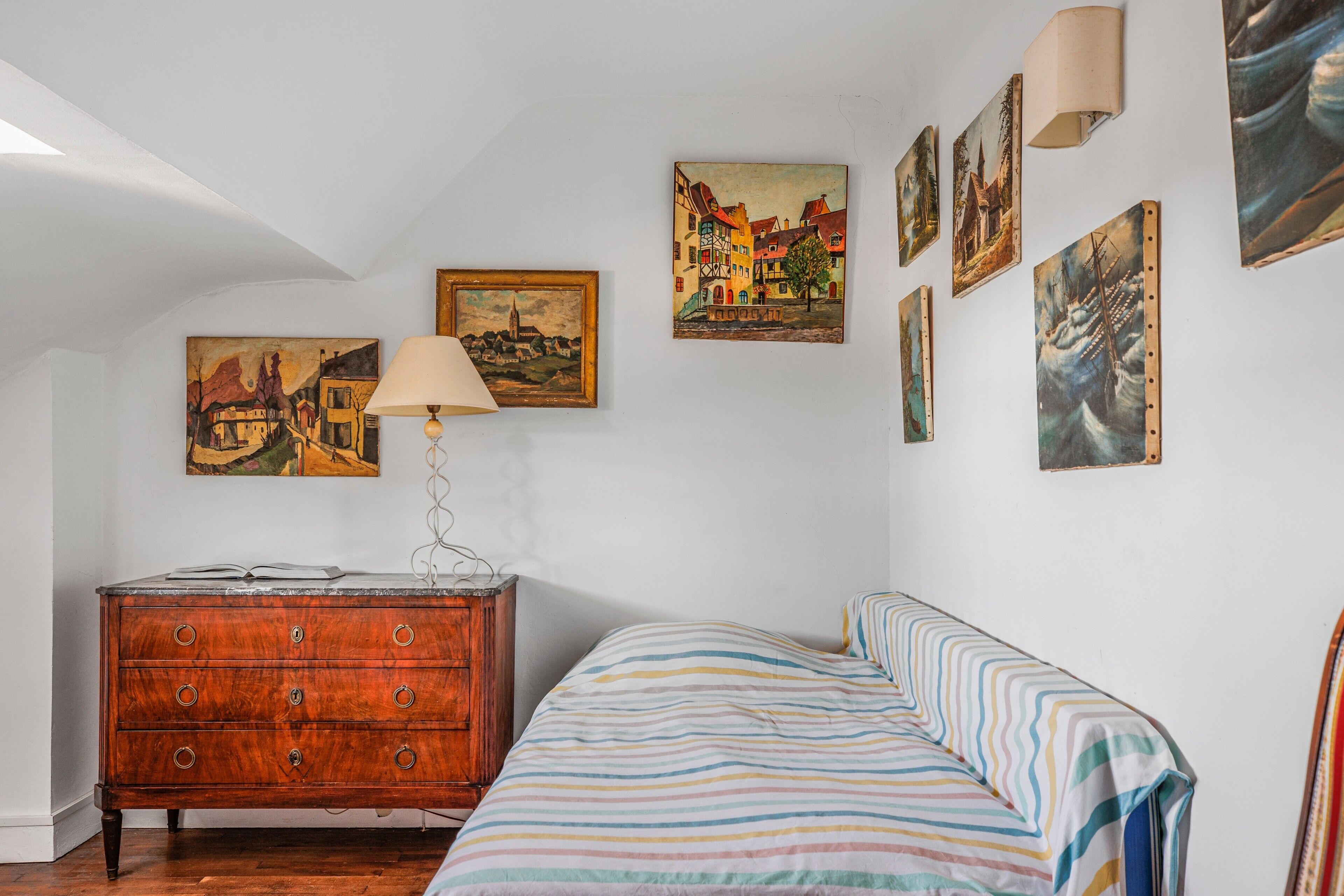 Une chambre avec une commode en bois, un lit avec une literie à rayures et plusieurs tableaux accrochés au mur.