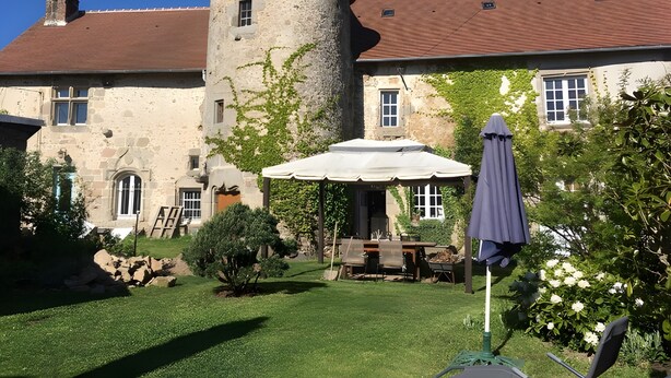 Un bâtiment en pierre doté d’une tour, une terrasse avec une table et des chaises, et un jardin avec des arbres et des fleurs.
