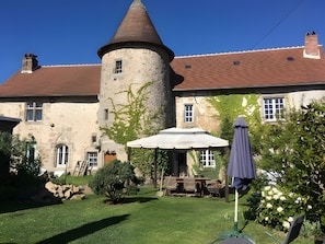 A stone building with a tower, a patio with a table and chairs, and a garden with trees and flowers.