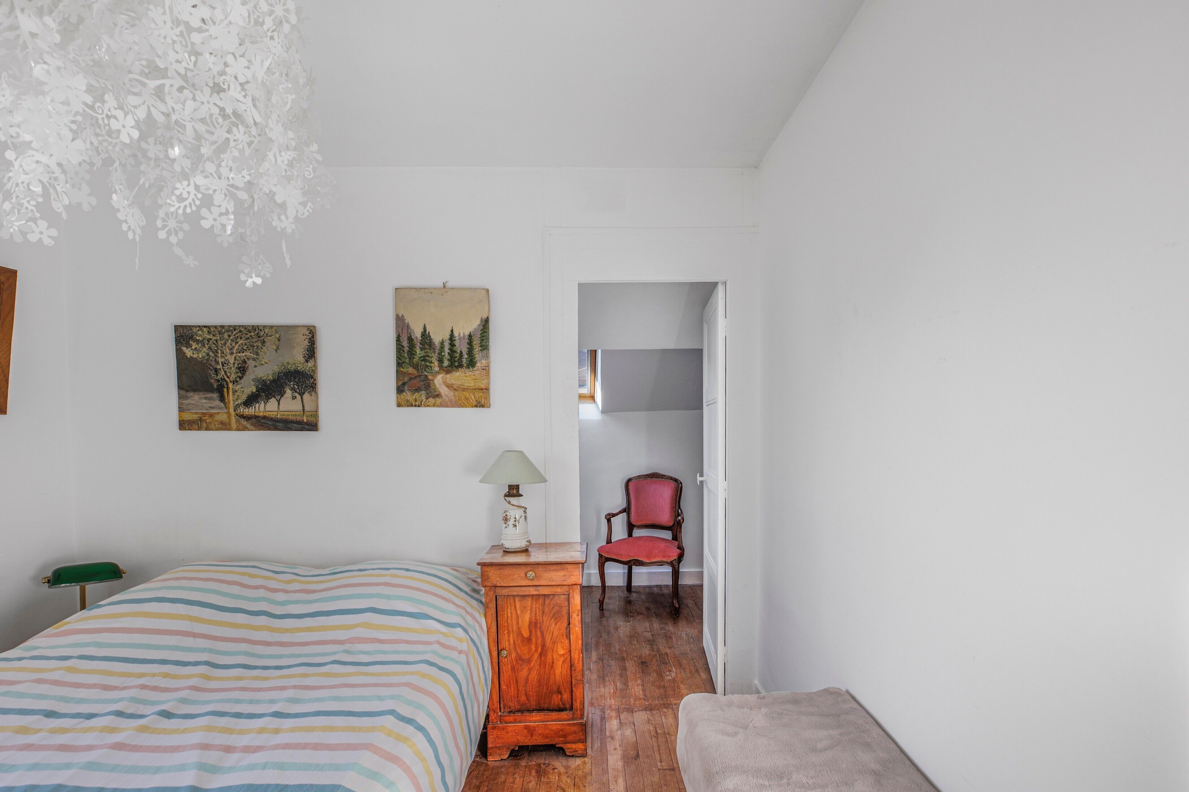 Una camera da letto con un letto, un comodino in legno, una sedia e una porta che dà su un'altra stanza.