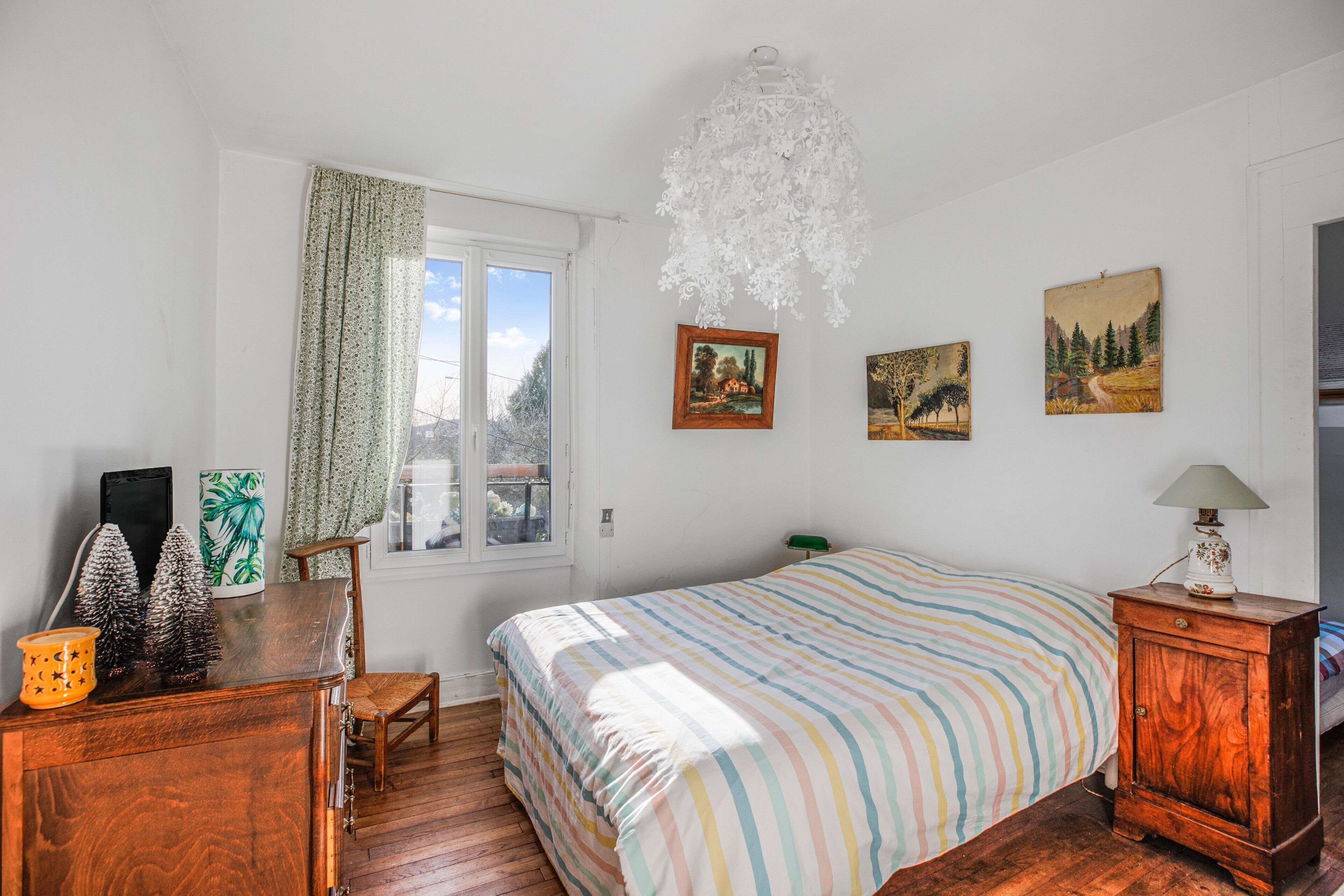 Ein Schlafzimmer mit Bett, einem Holzkleiderschrank, einem Schreibtisch, einem Stuhl, einem Fenster mit Vorhängen und Wandbildern.