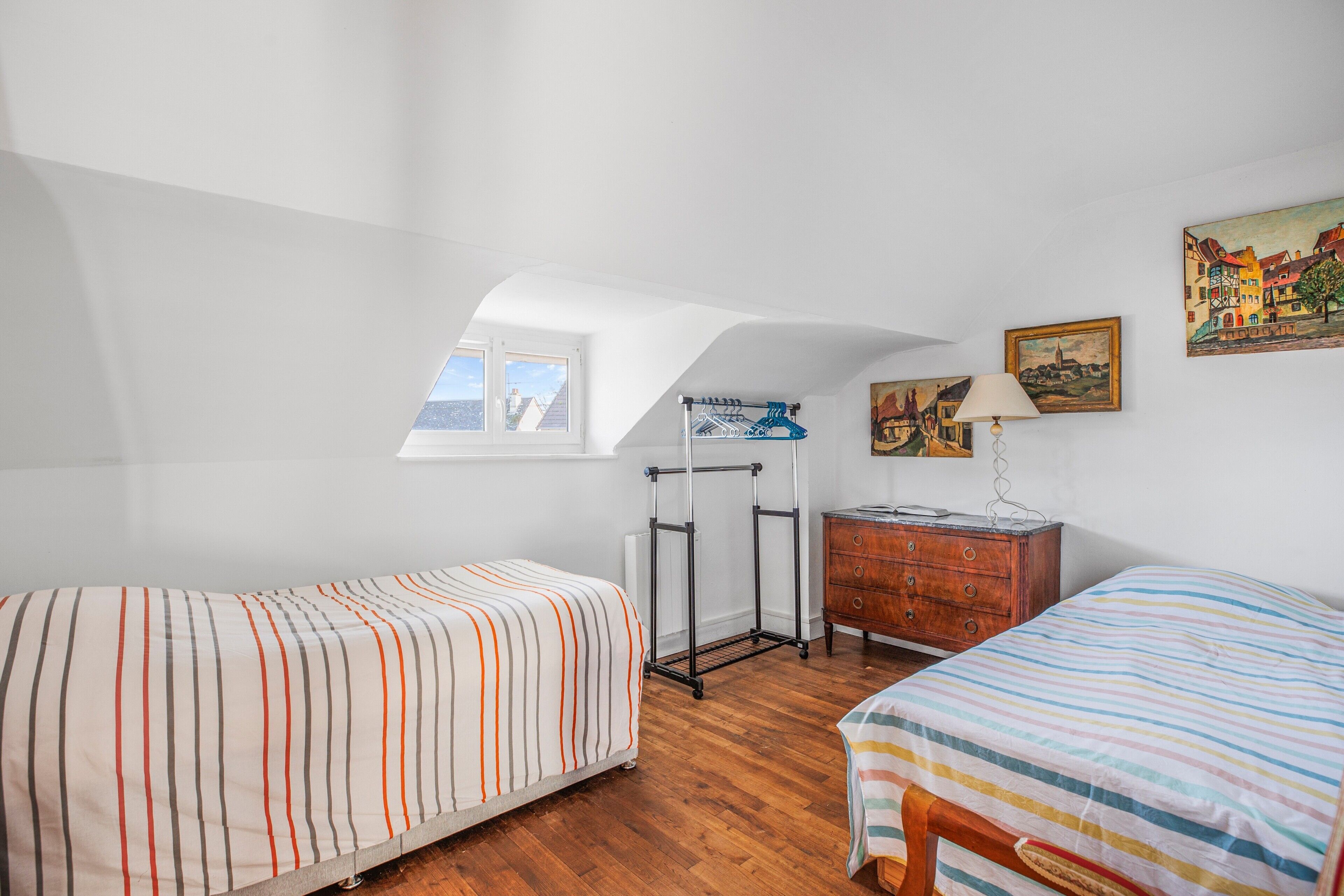 Quarto com duas camas, cómoda de madeira, candeeiro e um quadro na parede.