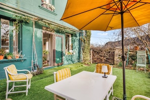 A patio with a white table and chairs, a yellow umbrella, and a green house. - Holiday Home 'Chez Poton' with Private Terrace, Shared Garden and Wi-Fi (Le Grand-Bourg)