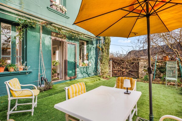 A patio with a white table and chairs, a yellow umbrella, and a green house.